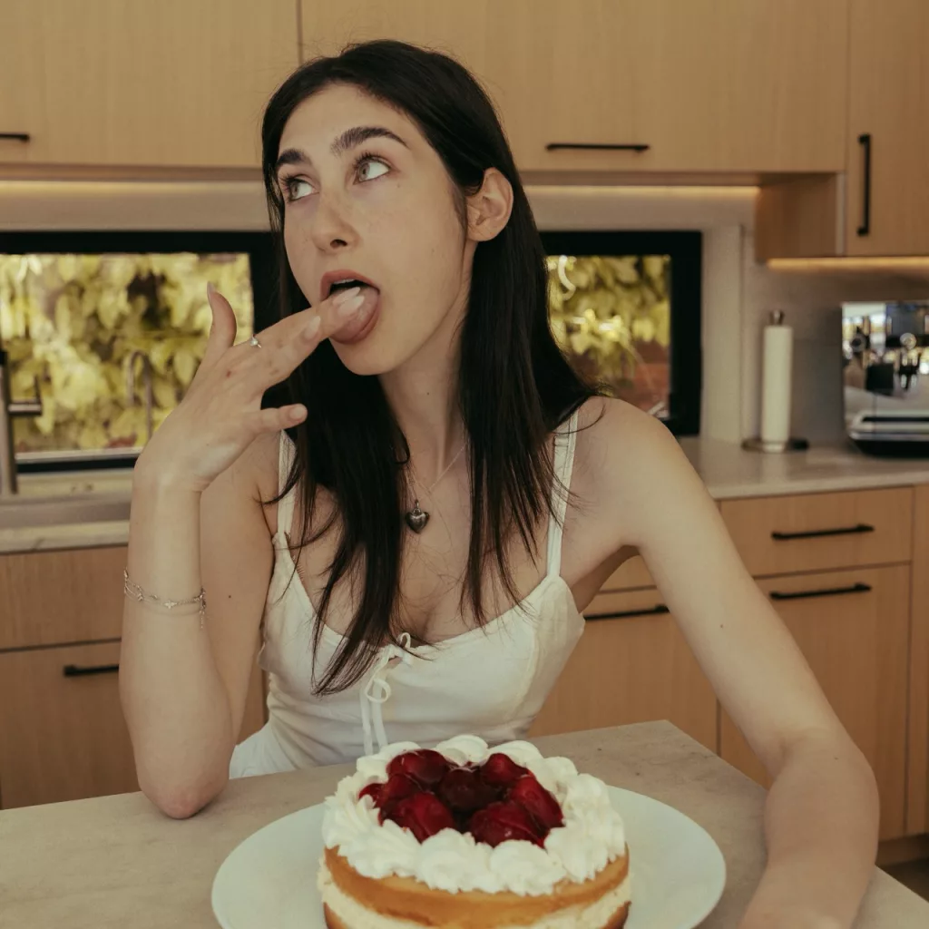 Yasmin Sydney in a kitchen, playfully licking frosting off her finger with a cherry-topped cake in front of her.