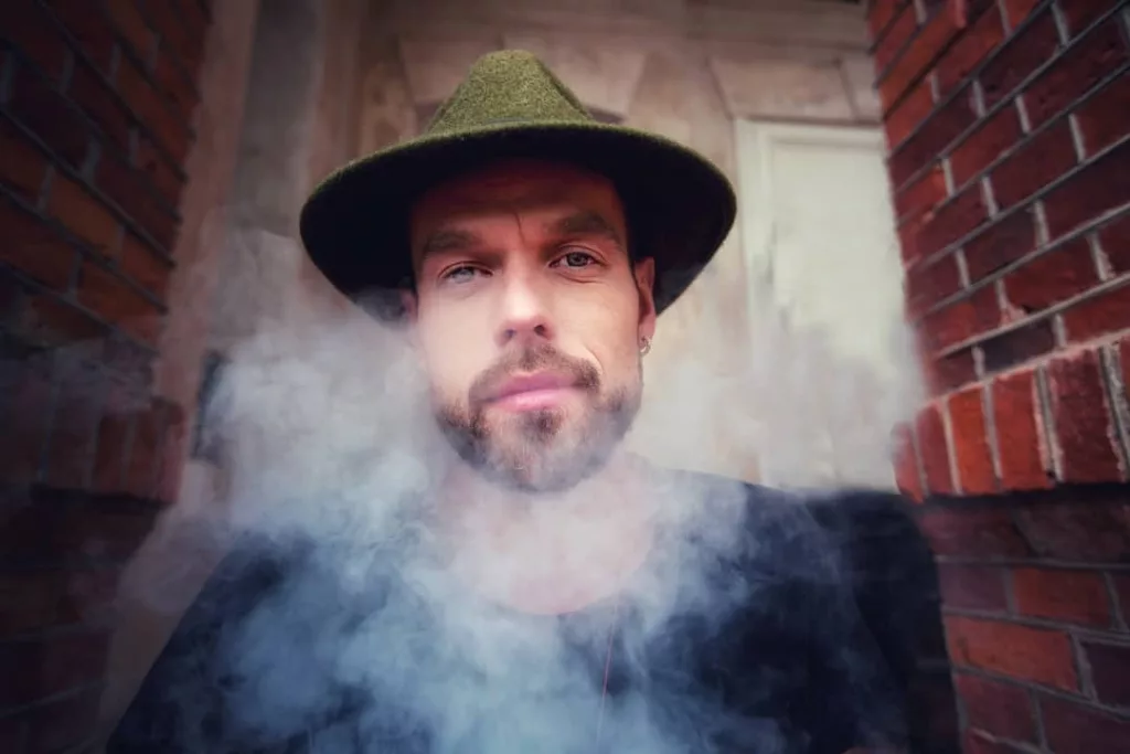 A bearded man with blue eyes and a contemplative expression wears a green hat and black shirt, surrounded by a cloud of smoke. He stands between two red brick walls, with a blurred background of a wooden door.