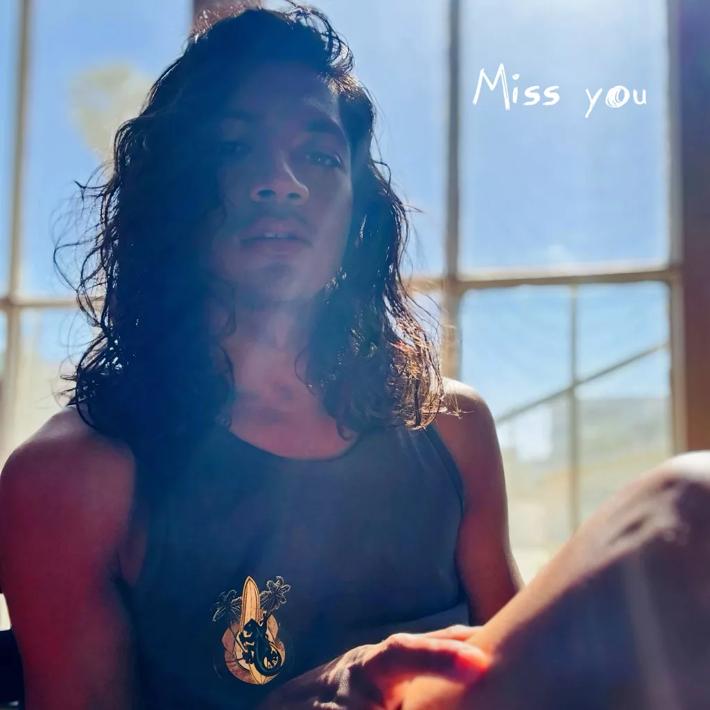 Bernard Shay Gomez sits in soft sunlight by a window, wearing a dark tank top, with the text "Miss you" appearing in white script in the top right corner.
