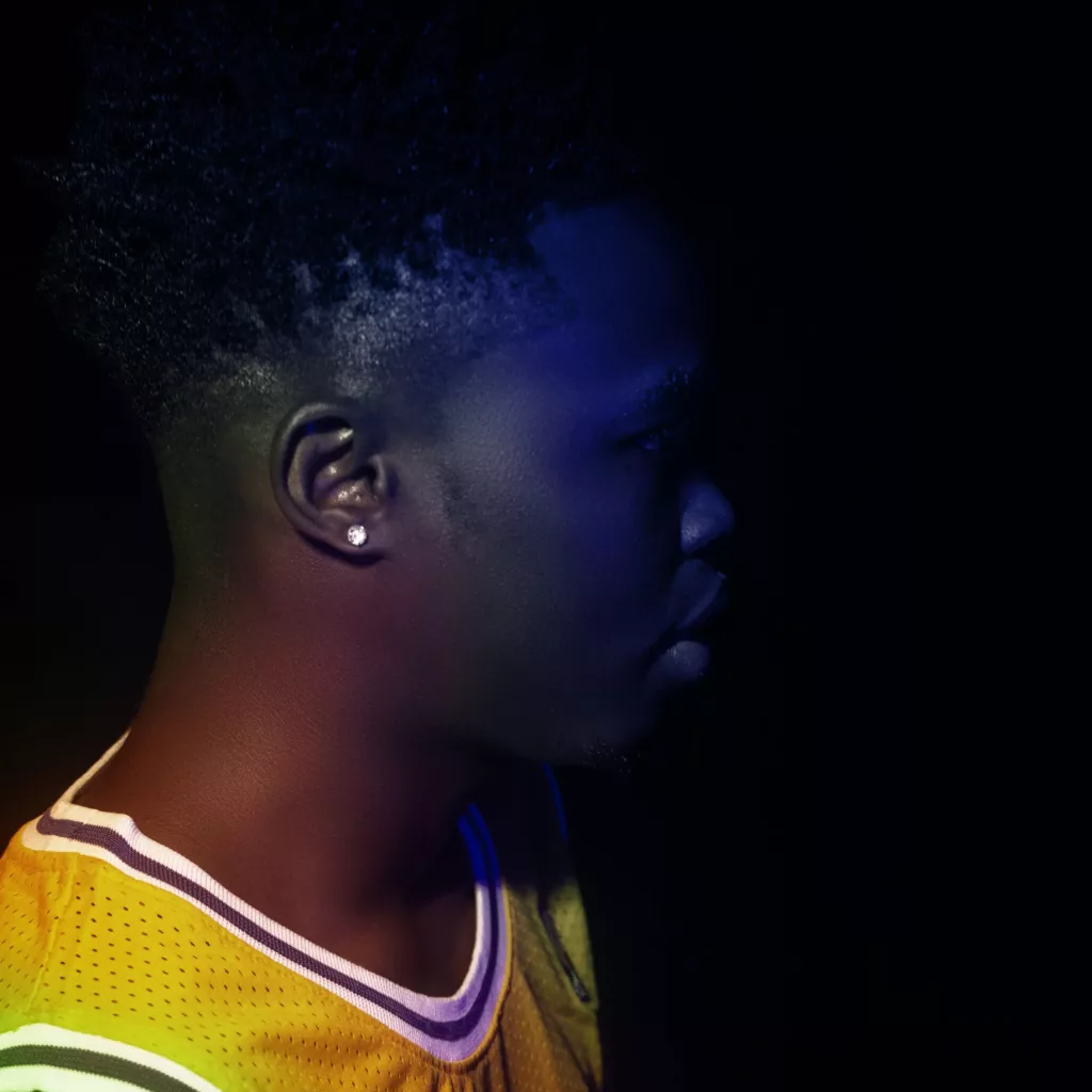 Profile image of Trapboi Sn wearing a yellow basketball jersey in a dimly lit setting with vibrant lighting effects.
