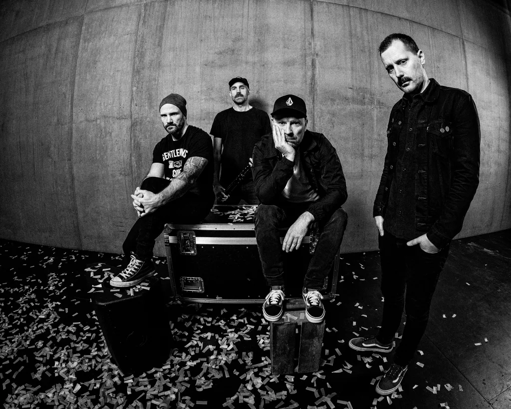 A black-and-white photo of the band Armatage Shanks, featuring four members posing against a concrete wall with confetti scattered on the ground, exuding a raw and punk aesthetic.