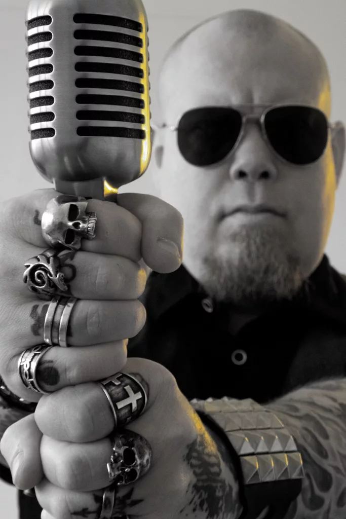 Black and white image of Fredrik Keith Croona, founder of Terrorbit, holding a vintage microphone. He is wearing sunglasses and multiple rings, exuding a bold and edgy persona.
