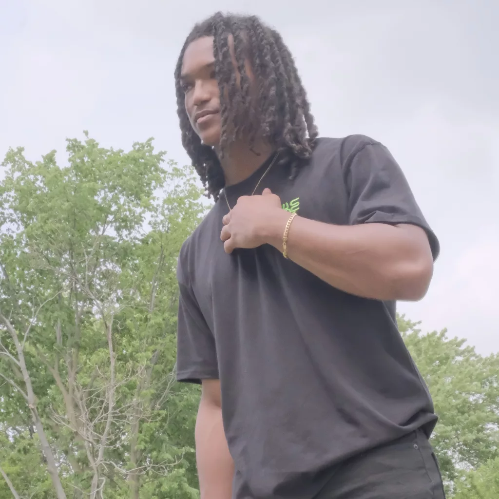 K. Bo24 in a black t-shirt standing outdoors, looking thoughtful with a hand on his chest.