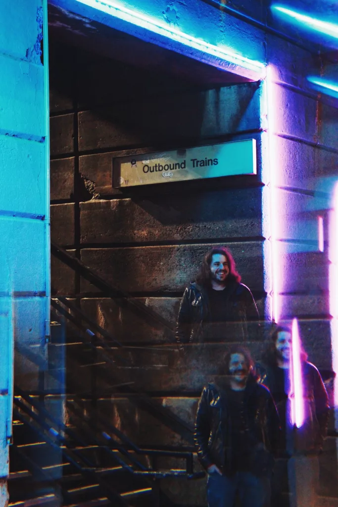 The image features Carter Fox standing near a dimly lit urban structure with a sign reading "Outbound Trains." The setting exudes a retro-futuristic vibe, accentuated by neon lights and dark blue tones. Carter is dressed in a leather jacket, blending into the urban, slightly gritty aesthetic. The reflection on the glass in front of him creates a double image, adding a sense of depth and complexity. This visual portrayal complements Carter's music style, which often explores chill-fi, jazz, and cosmic themes, suggesting a journey or exploration motif.