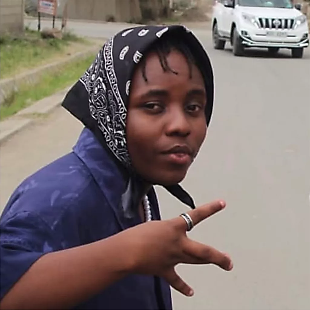 Young artist in a blue outfit and bandana making a hand gesture on a street, showcasing urban music culture.