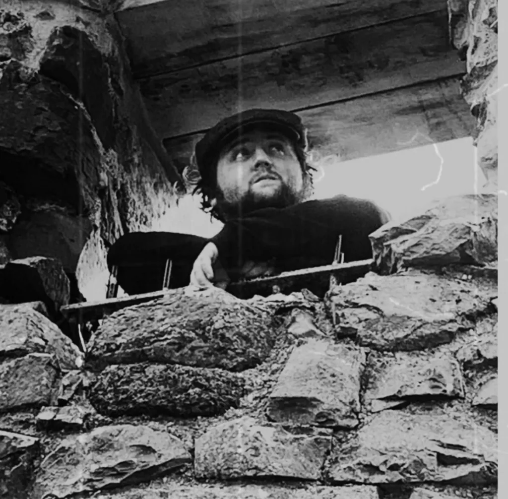 James Wilson peering through the stone ruins, wearing a flat cap and coat, evoking a rustic and folk-inspired aesthetic.
