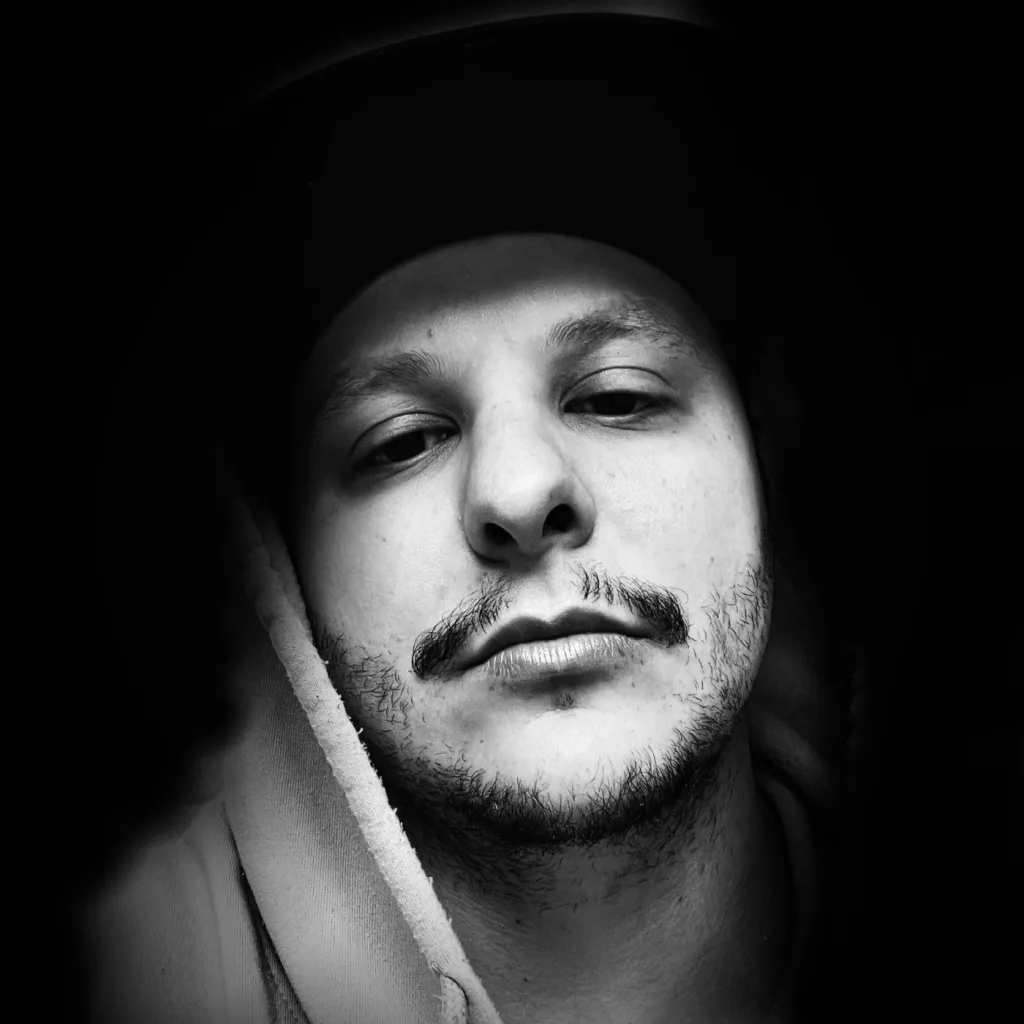 Black and white portrait of The Real J.T.W. featuring a close-up of the artist with a contemplative expression, wearing a hoodie and a cap.