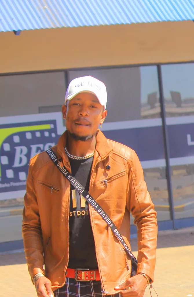 Geany-K, a rising hip-hop and Amapiano artist, wearing a brown leather jacket and white cap, posing confidently in front of a modern building.
