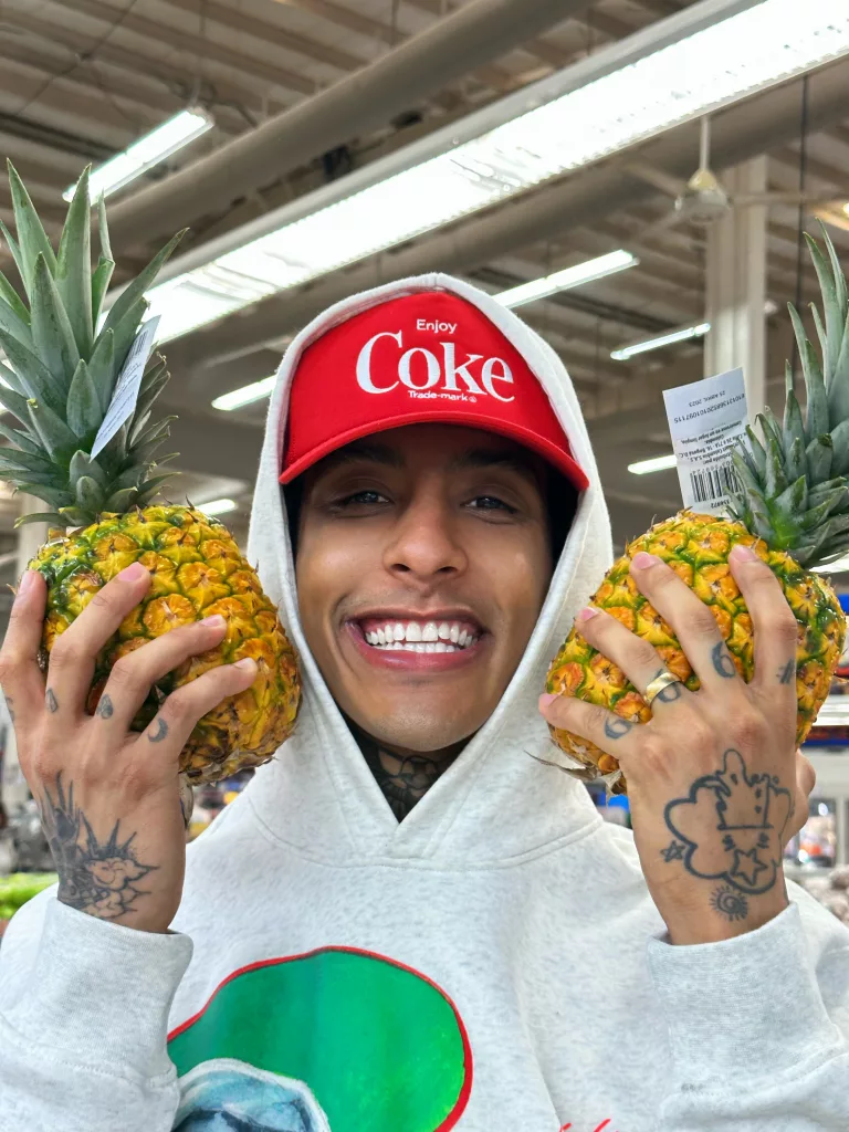 Till High, a smiling Colombian artist, holding two pineapples in a grocery store, wearing a white hoodie and a red cap with 'Enjoy Coke' written on it, showcasing his tattoos and vibrant personality.
