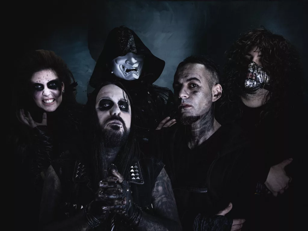 A dark, moody portrait of the industrial metal band TAZER. The five band members, dressed in gothic and industrial-style outfits, display intense expressions and dramatic face paint, set against a shadowy background.